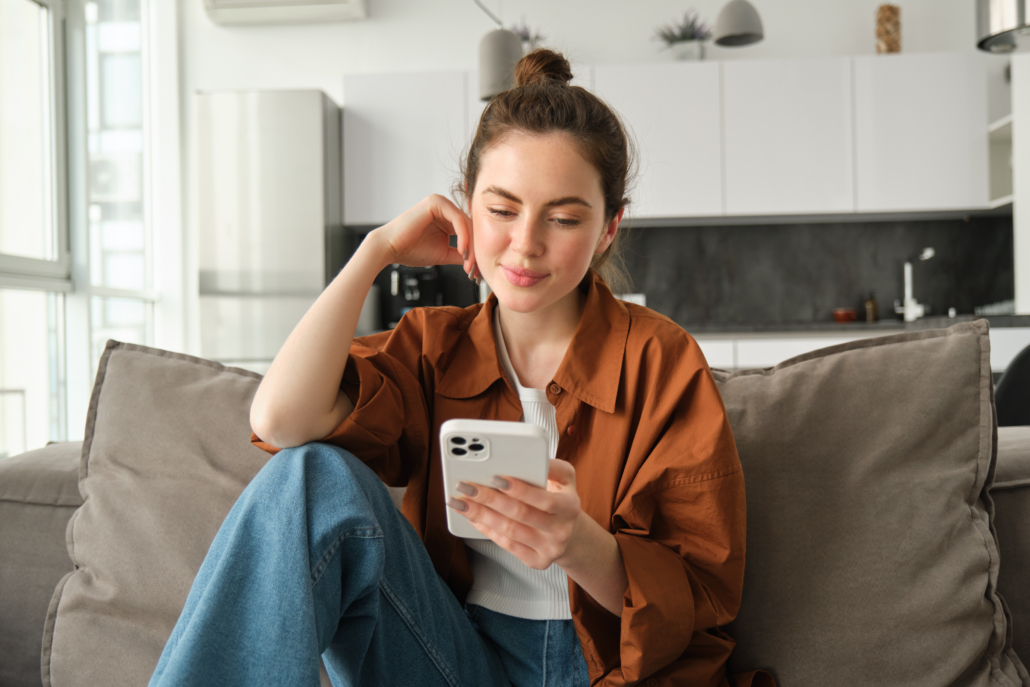 Portrait of happy young brunette woman sitting on sofa with smartphone, reading notification, messaging on mobile phone, resting on couch at home, smiling while looking at device screen